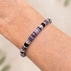 May include: A beaded bracelet featuring a repeating pattern of flat, circular purple and white striped beads, interspersed with glossy black cylindrical beads and small, metallic silver-toned spacer rings, arranged in a continuous circular band.