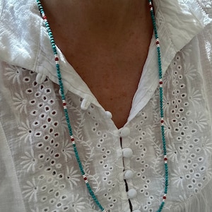 May include: A turquoise and red beaded necklace with silver accents. The necklace is worn by a person wearing a white blouse with an embroidered pattern.
