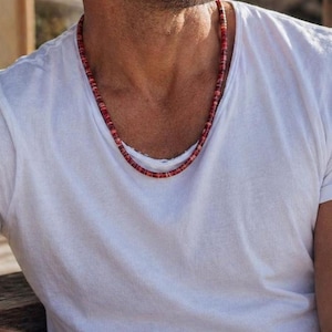 May include: A red and brown beaded necklace worn over a white t-shirt. The necklace is made of small, round beads. The t-shirt has rolled-up sleeves.
