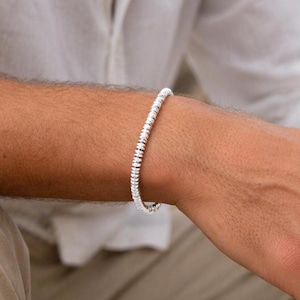 May include: A silver beaded bracelet is worn on a person's wrist. The bracelet is made of small, round beads strung together to form a flexible band. The beads have a shiny, metallic appearance.