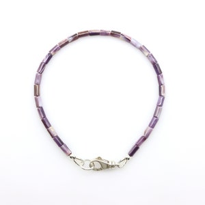 May include: A delicate bracelet featuring small, rectangular beads in shades of purple and lavender. The beads are strung together, forming a flexible chain. A silver-colored clasp secures the bracelet. The bracelet is displayed against a white background.
