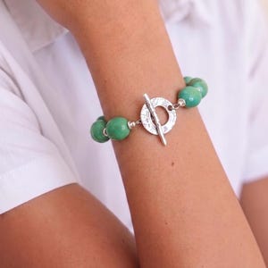 May include: A beaded bracelet featuring round, polished green stones interspersed with small silver-toned beads. The bracelet is secured with a circular, textured silver-toned toggle clasp that has a bar-shaped closure piece.