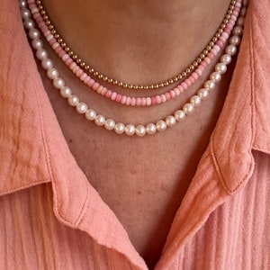 May include: A layered necklace with three strands of beads. The top strand is gold, the middle strand is pink, and the bottom strand is white.