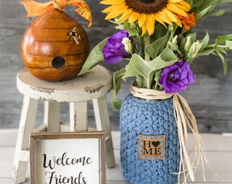 May include: A rustic, decorative scene featuring a wooden beehive, a small white stool, a vase with a blue knitted cover, and a framed sign that reads "Welcome Friends." The vase holds a vibrant bouquet of sunflowers and purple flowers.