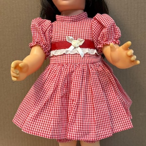 Peut inclure: Une poupée aux cheveux bruns et aux yeux bleus portant une robe à carreaux vichy rouge et blanc avec un bonnet assorti. La robe présente une ceinture rouge avec un nœud en ruban blanc et une bordure en dentelle. Chaussettes et chaussures rouges.