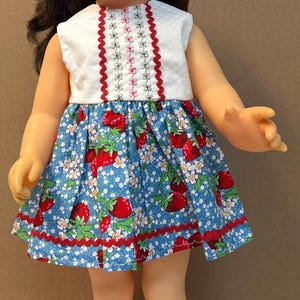 Op de afbeelding: Een pop met donkerbruin haar en blauwe ogen draagt een wit mouwloos topje met rode zigzagband en een blauwe rok met een patroon van rode aardbeien en witte bloemen, afgewerkt met een rode zigzagband aan de zoom en witte sandalen.