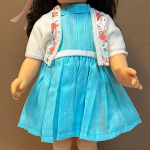 Peut inclure: Une poupée aux cheveux brun foncé et aux yeux bleus portant une robe turquoise vif avec un empiècement blanc, un gilet blanc à manches courtes avec bordure florale, des chaussettes blanches à dentelle et des chaussures à fleurs roses.