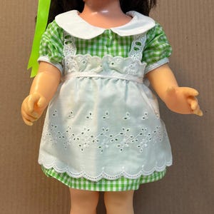 Op de afbeelding: Een pop met donkerbruin haar, blauwe ogen en sproeten, gekleed in een groen-wit geruite jurk met een wit schortje met broderie anglaise, een witte kraag, witte sokken en lichtgroene schoenen. Een felgroen lint in het haar.
