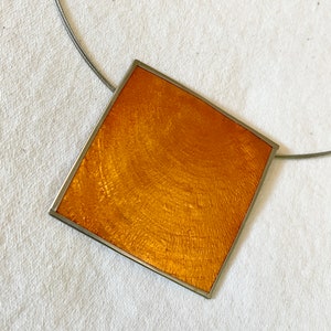 May include: A gold square pendant with a textured surface, hanging from a silver chain.