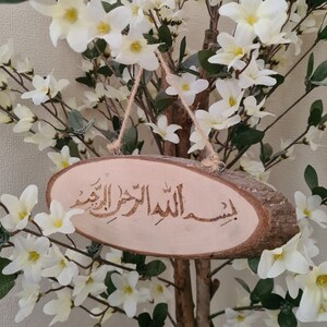 May include: A wooden sign with the Arabic phrase "Insha'Allah" carved into it, hanging from a branch of a white flowering tree.