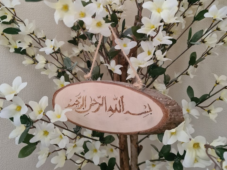 May include: A wooden sign with the Arabic phrase "Insha'Allah" carved into it, hanging from a branch of a white flowering tree.