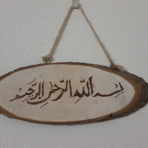 May include: A wooden sign with a brown rope hanging from the top. The sign has the Arabic phrase "Bismillah al-Rahman al-Rahim" written on it.