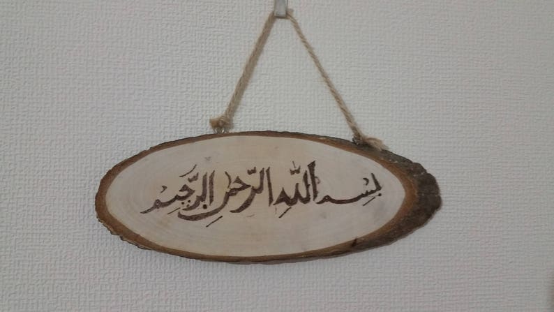 May include: A wooden sign with a brown rope hanging from the top. The sign has the Arabic phrase "Bismillah al-Rahman al-Rahim" written on it.