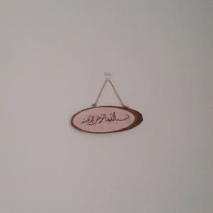 May include: A wooden oval sign with the Arabic phrase "Bismillah al-Rahman al-Rahim" written in calligraphy. The sign is hanging on a white wall with a string.