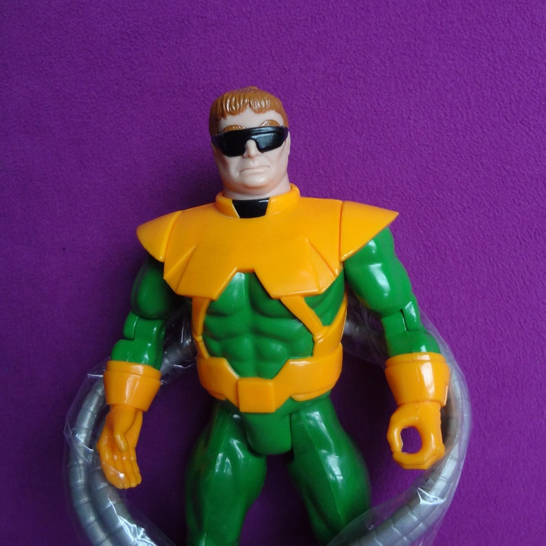 May include: A green and yellow action figure of a superhero wearing a mask and glasses. The figure has four mechanical arms.