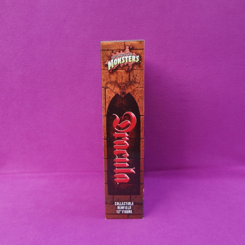 May include: A rectangular box featuring the Universal Studios Monsters logo and the name "Dracula" in red script. The box is brown with a brick wall design and a bat graphic. The text "COLLECTIBLE RENFIELD 12" FIGURE" is at the bottom.