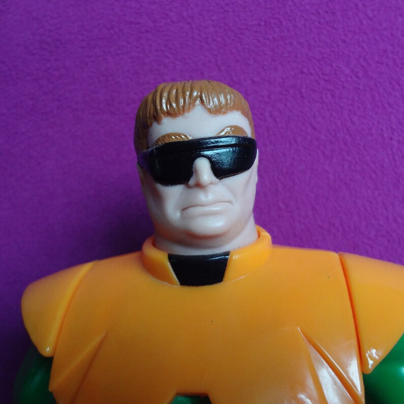 May include: A plastic action figure of a man with brown hair wearing black sunglasses and an orange and green costume.