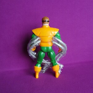 May include: A green and orange action figure of Doctor Octopus, a supervillain from the Marvel Comics universe. The figure has four mechanical arms and is posed with its back to the camera.