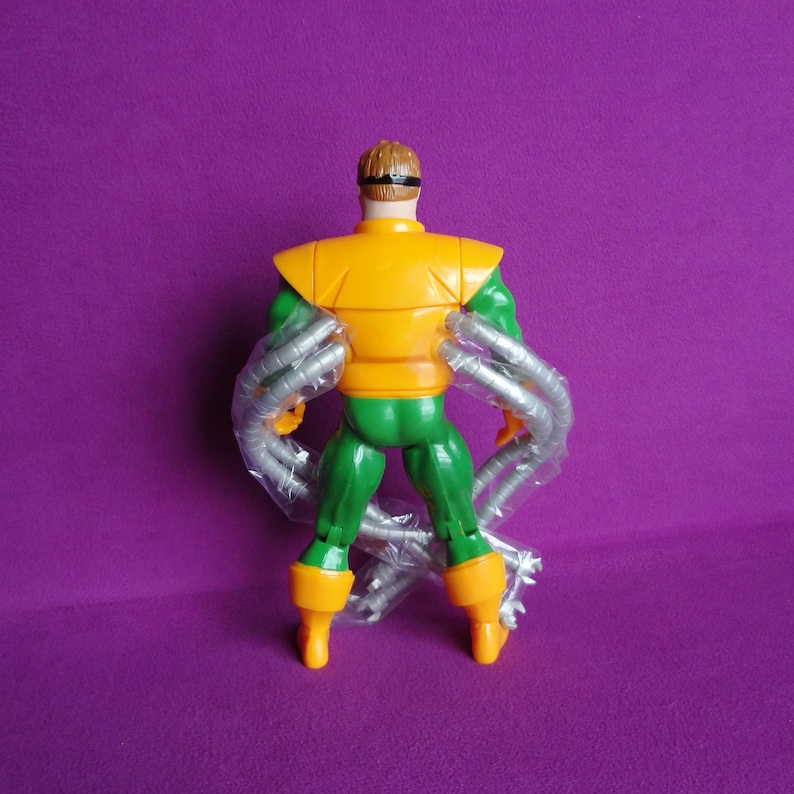 May include: A green and orange action figure of Doctor Octopus, a supervillain from the Marvel Comics universe. The figure has four mechanical arms and is posed with its back to the camera.