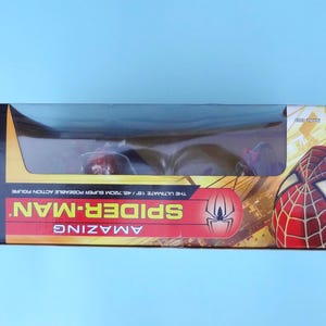 Puede incluir: Una figura de acci&oacute;n de Spider-Man en una caja. La caja es roja, amarilla y negra, con las palabras "Spider-Man" y "Amazing" en un lugar destacado. La figura es visible a trav&eacute;s de una ventana de pl&aacute;stico transparente. La caja mide 45,72 cm.