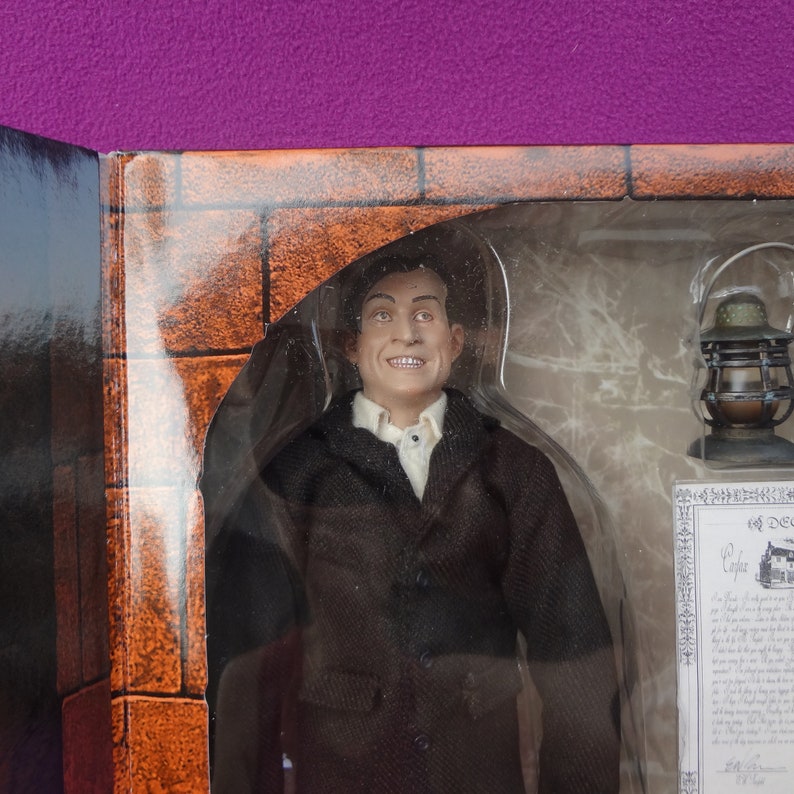May include: A figure of a man in a black suit and white shirt, smiling, inside a clear plastic case. A small lantern and a document are also visible. The background is a brick wall design.