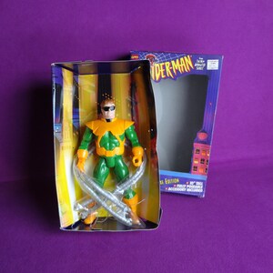 May include: A green and orange action figure of Doc Ock from the Spider-Man animated series. The figure is packaged in a box with a cityscape background. The box features the text "Spider-Man" and "Deluxe Edition".