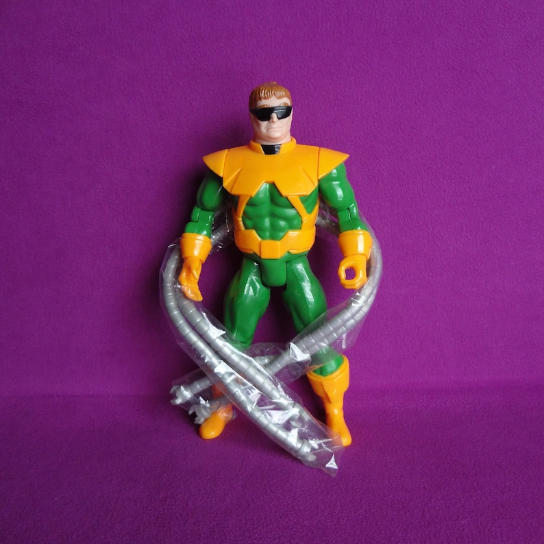 May include: A green and orange action figure of Doctor Octopus, a supervillain from the Spider-Man comics. The figure has four mechanical arms and is wearing a mask and goggles.