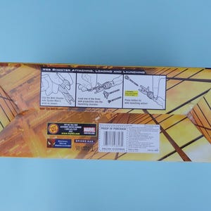 Puede incluir: Una caja rectangular con un dise&ntilde;o geom&eacute;trico amarillo y marr&oacute;n. La caja presenta ilustraciones y texto que detallan el uso de un lanzador de telara&ntilde;as. El texto incluye "WEB SHOOTER ATTACHING, LOADING AND LAUNCHING" y "SPIDER MAN".