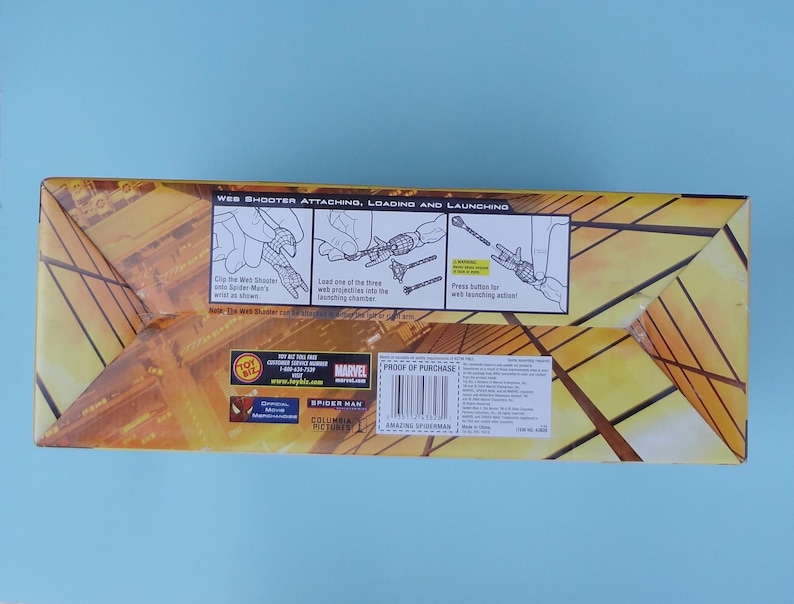 Puede incluir: Una caja rectangular con un dise&ntilde;o geom&eacute;trico amarillo y marr&oacute;n. La caja presenta ilustraciones y texto que detallan el uso de un lanzador de telara&ntilde;as. El texto incluye "WEB SHOOTER ATTACHING, LOADING AND LAUNCHING" y "SPIDER MAN".