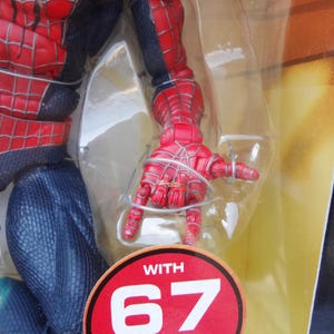 Puede incluir: Una figura de acci&oacute;n de Spider-Man roja y negra, parcialmente visible, en un embalaje de pl&aacute;stico transparente. La mano de la figura est&aacute; extendida, con detalles de telara&ntilde;a. Una pegatina circular roja y blanca dice "WITH 67".