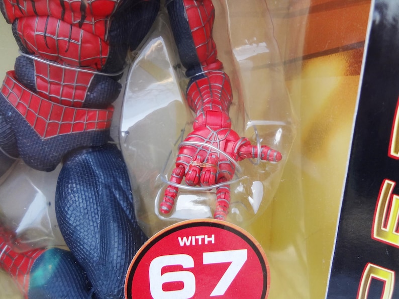 Puede incluir: Una figura de acci&oacute;n de Spider-Man roja y negra, parcialmente visible, en un embalaje de pl&aacute;stico transparente. La mano de la figura est&aacute; extendida, con detalles de telara&ntilde;a. Una pegatina circular roja y blanca dice "WITH 67".