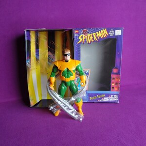 May include: A green and yellow action figure of Doctor Octopus from the Spider-Man animated series. The figure is holding its four tentacles and is posed in a fighting stance. The figure is packaged in a box with the Spider-Man logo and the words "Deluxe Edition".