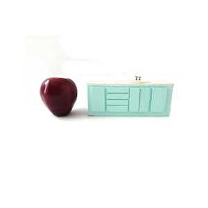 May include: A miniature, light blue wooden cabinet with a light-colored countertop. The cabinet has three drawers and two doors. A shiny red apple sits to the left of the cabinet. The cabinet is a decorative item.