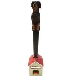 May include: A decorative pen featuring a Rottweiler dog figurine atop a dark brown shaft. The pen's base is a miniature dog house with a red roof and a small orange object visible inside. The dog is brown and black.