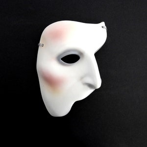 May include: A white half-face mask featuring a smooth, contoured surface with a prominent nose and eye opening. The mask displays subtle pinkish-red blush accents on the forehead and cheek area, with small grey elastic loops attached at the sides.