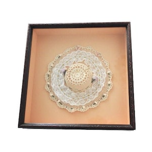 May include: A framed, vintage-style artwork featuring a cream-colored, crocheted hat and doilies. The hat is centered on a peach-colored background within a dark brown wooden frame. The doilies have a delicate, lacy design.