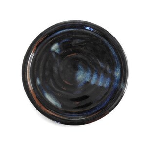 May include: A handmade ceramic plate with a dark, glossy finish. The plate features a swirling pattern of black, blue, and brown hues. The rim has a subtle brown accent. This unique piece is perfect for serving food or as a decorative item.