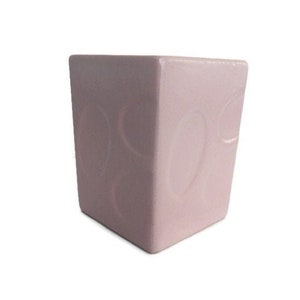 May include: A pale pink ceramic square container featuring a subtle embossed pattern of overlapping circles on its exterior surface. The object has a smooth, glossy finish and a clean, geometric shape with straight vertical sides and sharp corners.