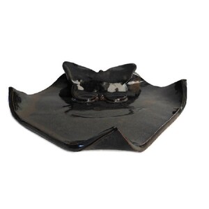 May include: A dark, glossy ceramic dish with a square-like shape and upturned edges. A sculpted butterfly figure is attached to the center of the dish. The surface has a reflective, dark brown and black glaze with subtle variations in color tone.