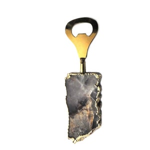 May include: A bottle opener with a gold-colored metal top and a natural stone handle. The stone is a mix of gray, brown, and white with gold edging. The opener is designed for opening bottles.