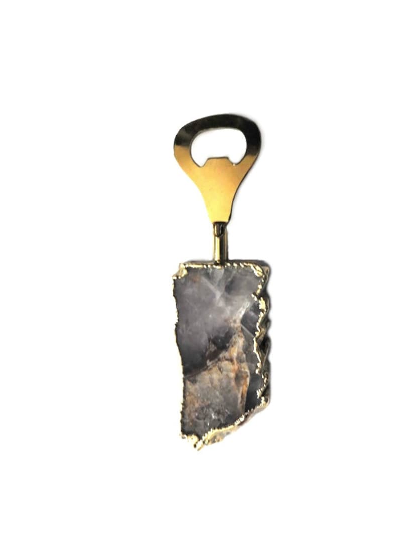 May include: A bottle opener with a gold-colored metal top and a natural stone handle. The stone is a mix of gray, brown, and white with gold edging. The opener is designed for opening bottles.