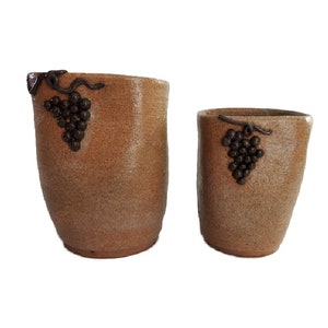 May include: Two brown ceramic vases with a textured surface. Each vase features a dark brown grape cluster design. The vases are different sizes, with a taller, wider vase and a smaller, narrower vase.