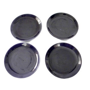 May include: Four dark blue ceramic plates arranged in a square formation. Each plate has a slightly raised rim and a smooth, glossy surface. The plates are round and appear to be of uniform size, suitable for serving appetizers or desserts.