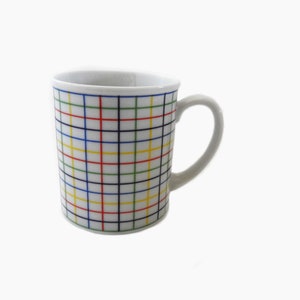 May include: A white ceramic mug with a colorful grid pattern. The grid is composed of intersecting lines in red, yellow, green, blue, and black. The mug has a curved handle and a simple, clean design.