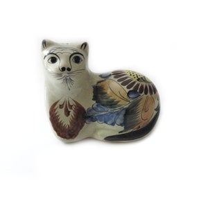 May include: Ceramic cat figurine with a hand-painted floral design. The cat is primarily off-white with black facial details and a brown, blue, and yellow floral pattern on its back. A decorative piece for cat lovers.