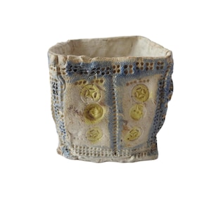May include: A handmade ceramic planter with a square shape. The exterior features a textured, off-white surface with blue and yellow accents. The design includes embossed circular patterns and a lace-like border.