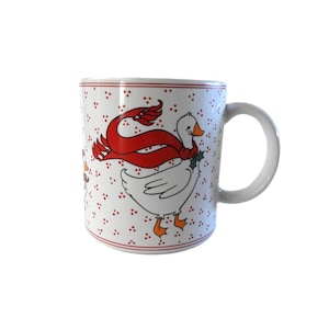 May include: White ceramic mug with a whimsical goose design. The goose wears a red scarf and is surrounded by red polka dots. The mug has a white handle and a red trim.