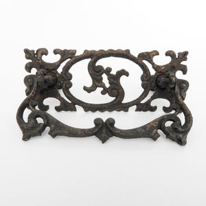 May include: Ornate, antique-style drawer pull with a dark bronze finish. The pull features an oval center design with scrolling details and decorative end pieces. A curved handle is below the oval.