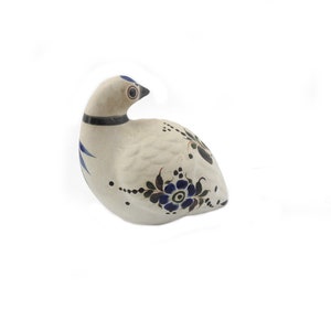 May include: A ceramic bird figurine in a resting pose. The bird is primarily off-white with blue and black floral and geometric designs. It has a black collar and a dark beak. The figurine is on a white background.