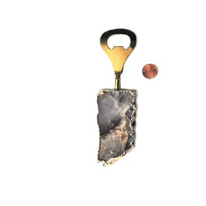 May include: A bottle opener with a brass finish and a natural stone handle. The stone is a mix of gray and purple with gold edging. A penny is next to the bottle opener for size comparison.